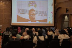 Foto suasana aula seminar saat perwakilan dari Kementerian Pemberdayaan Perempuan dan Perlindungan Anak (KemenPPPA) memberikan sambutan atau tanggapan secara daring melalui layar proyektor besar. Di depan panggung, para peserta seminar duduk menyimak pemaparan tersebut dari area audiens.